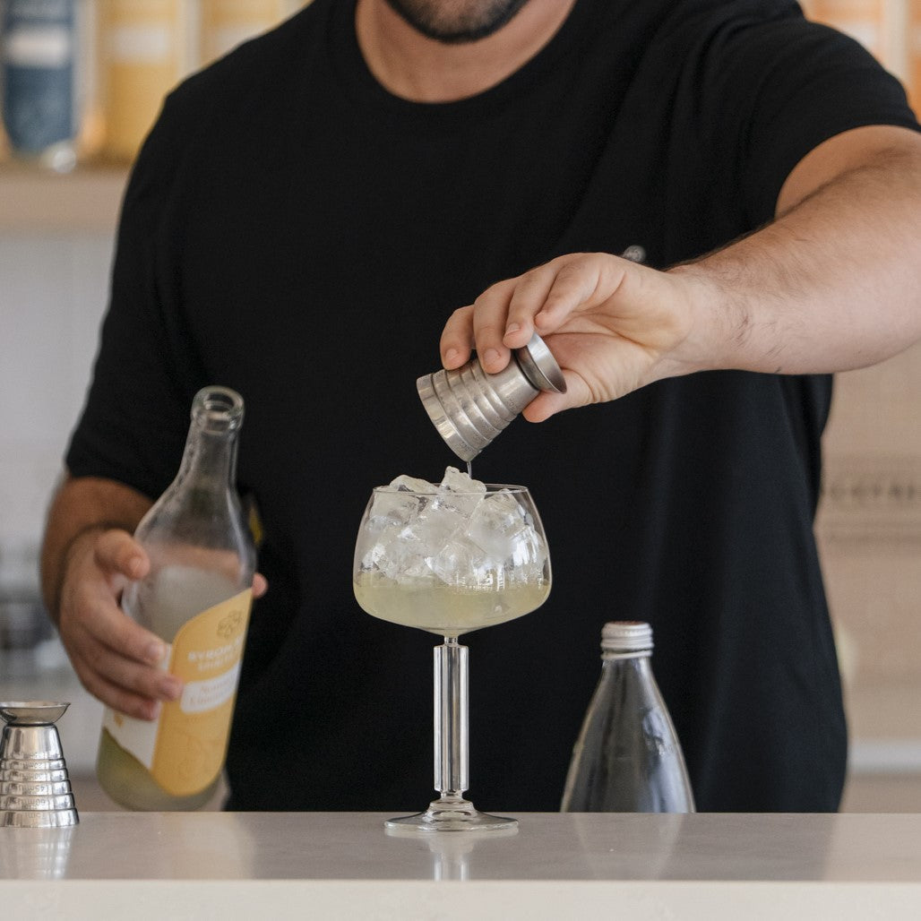 person pouring a shot of Limoncello into cocktail glass filled with ice, while holding open limoncello bottle. Background blurred