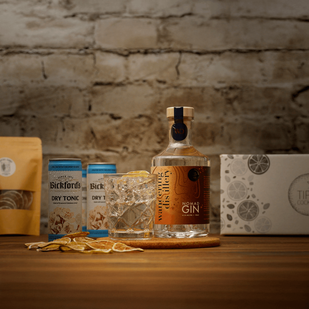 Bottle of Wandering Distillery Nomad Gin with tonic water cans on a wooden surface with a stone wall background.