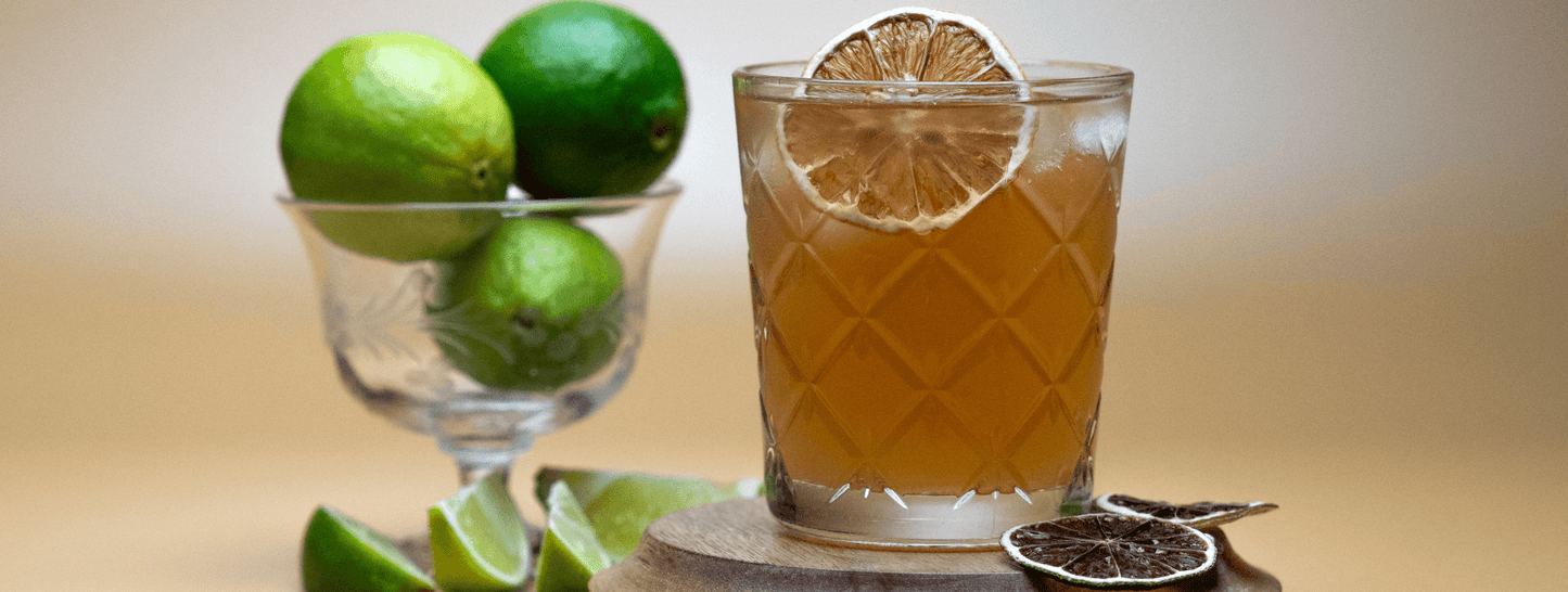 The Forecast Cocktail on a wooden coaster with dried lime garnish, next to a glass cup of fresh limes with a very light yellow background