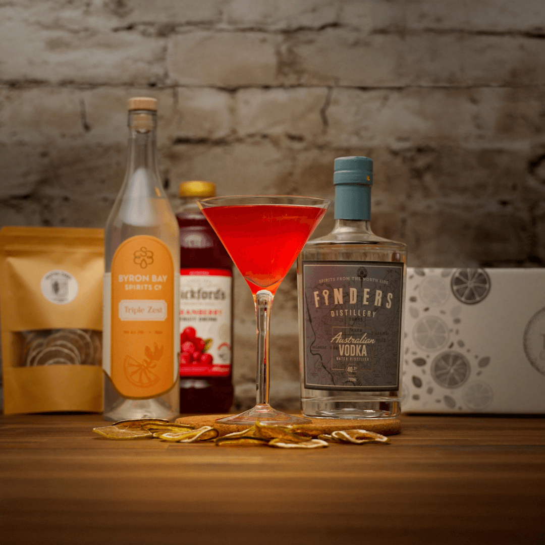 Bottles of Finders Vodka, a cocktail glass with pink cosmopolitan liquid, and other items on a wooden surface with a stone wall background.