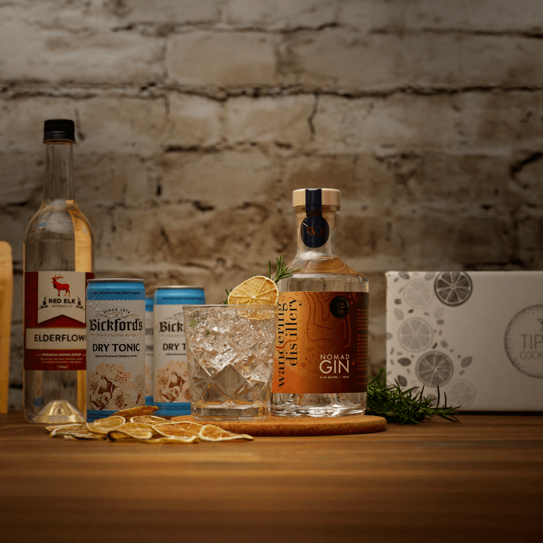 Bottles of Wandering Distillery Nomad Gin with tonic water and Red Elk Elderflower syrup with a cocktail on a wooden surface against a brick wall