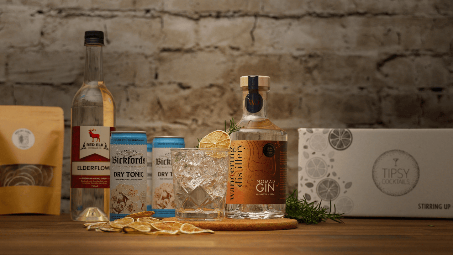 Cocktail kit showing cocktail with bottle of gin, elderflower syrup and other ingredients on wooden bench with stone background