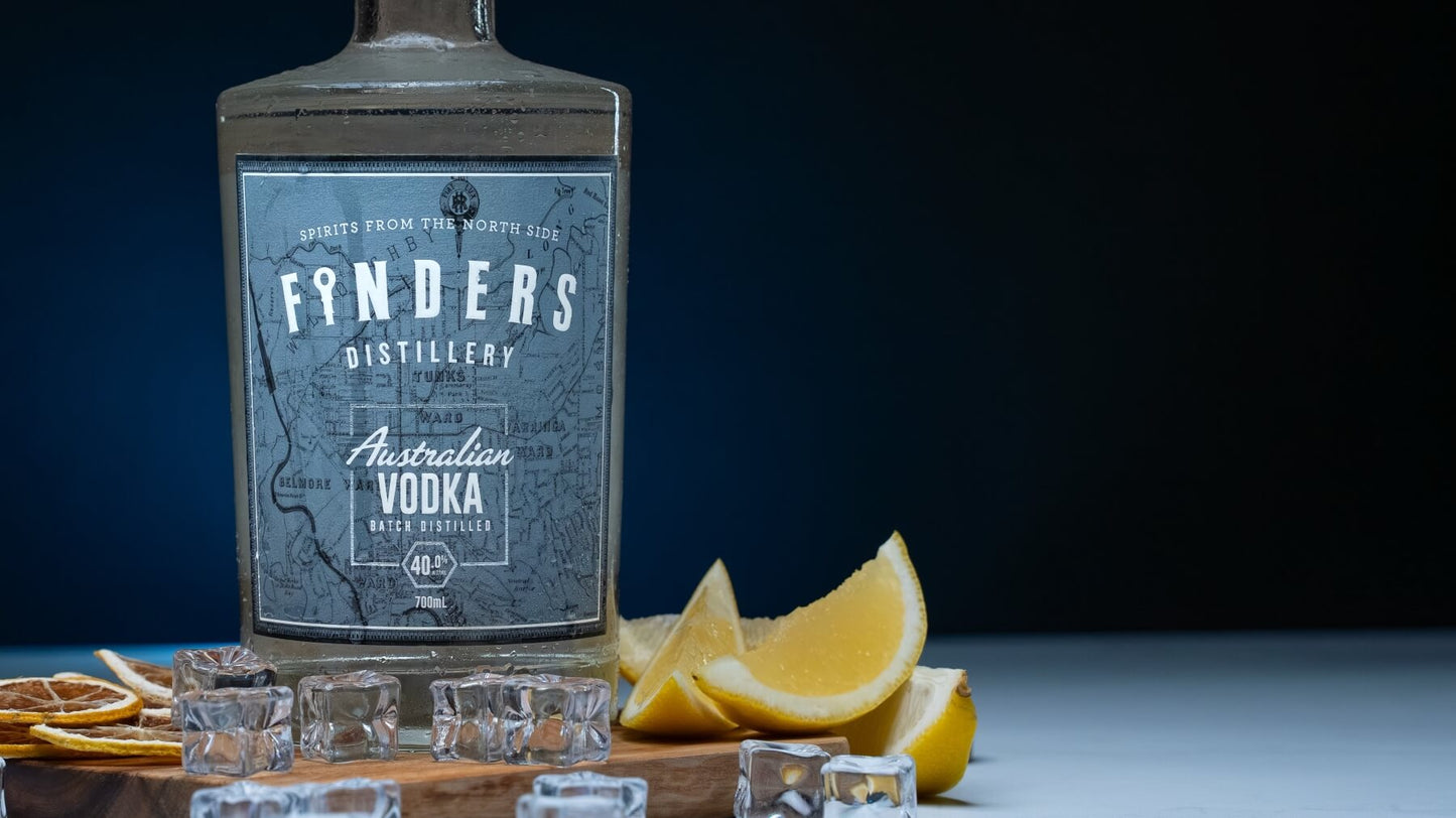 Finders Distillery Australian Vodka on a wooden board surrounded by fresh lemon wedges, ice cubes and dried lemon slices. With a dark blue background