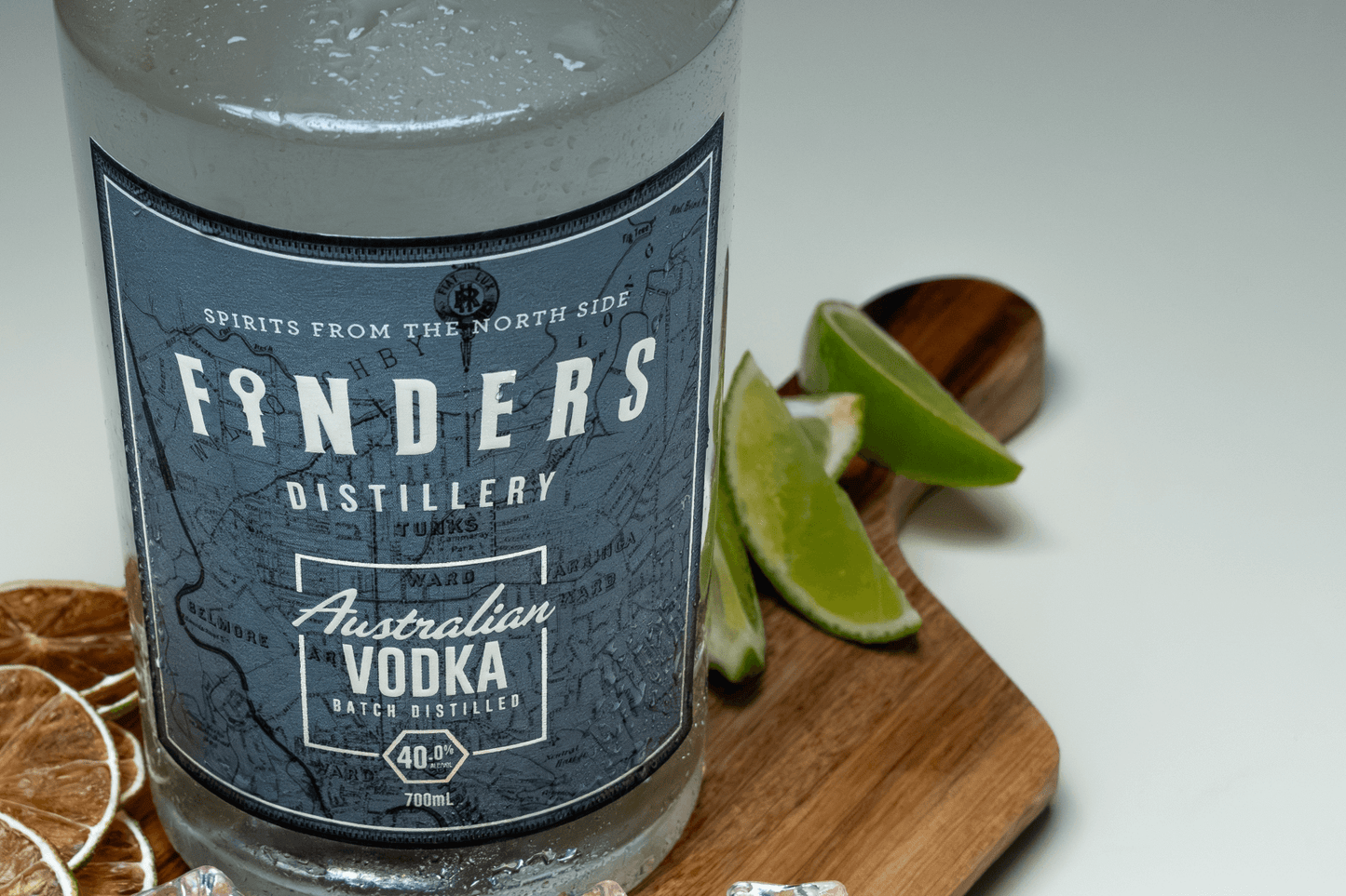 Finders Distillery Bottle Close Up on a wooden board with fresh lime on one side, and dried lime slices on the other side. Very light blue background