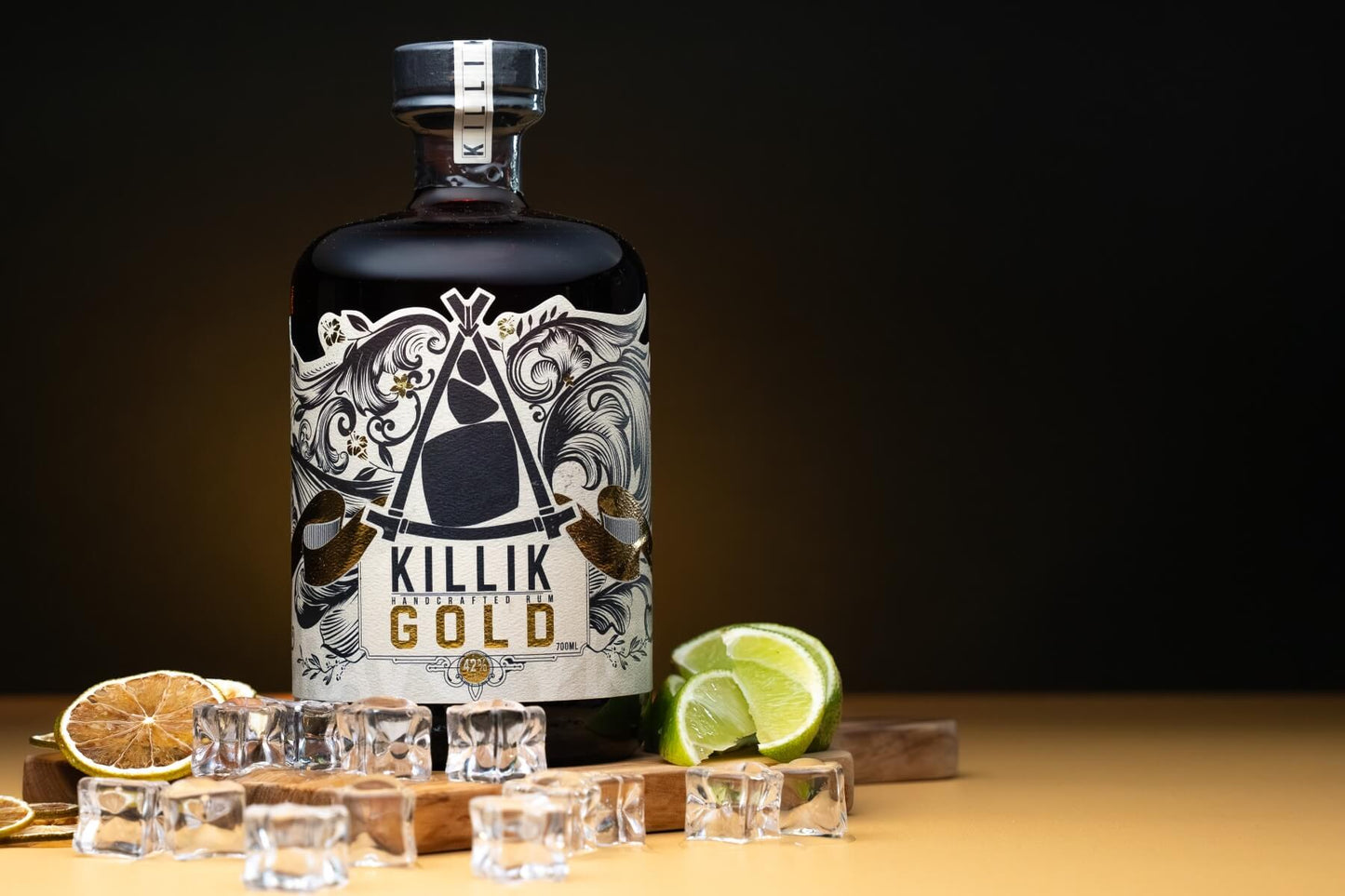 Bottle of Killik Gold on wooden board surrounded by ice, fresh lime and dried lime slices. Dark background with orange glow behind bottle.