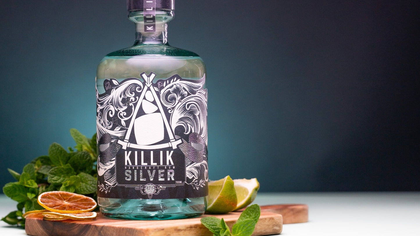 Bottle of Killik Silver Rum on a wooden board with fresh limes on one side of the bottle, dried lime slices on the other side of the bottle and a bunch of fresh mint leaves to the side and back. Dark Blue background