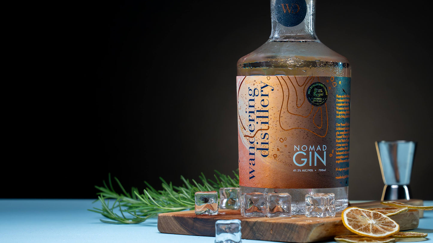 Bottle of Wandering Distillery Nomad Gin on a wooden board with ice, dried lemon and fresh rosemary in the background.