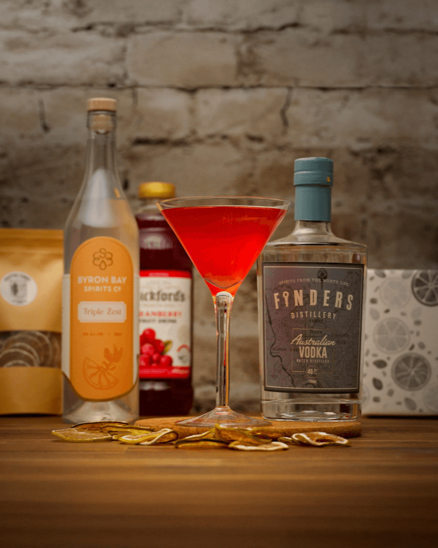 Beverage setup with bottles, a cocktail glass, and packaging on a wooden surface with a stone wall background.