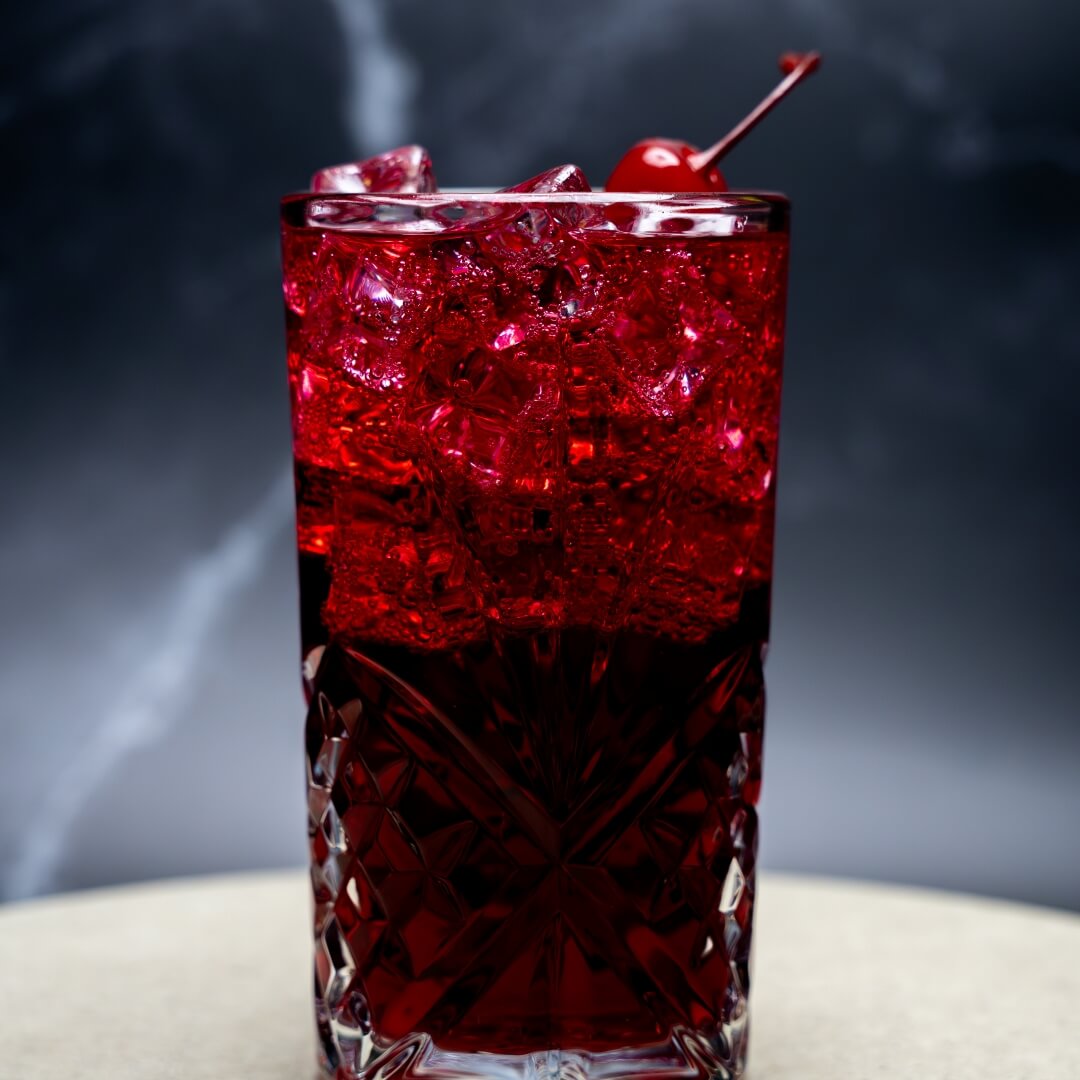 Red mocktail with ice and a cherry in a textured glass on a dark background