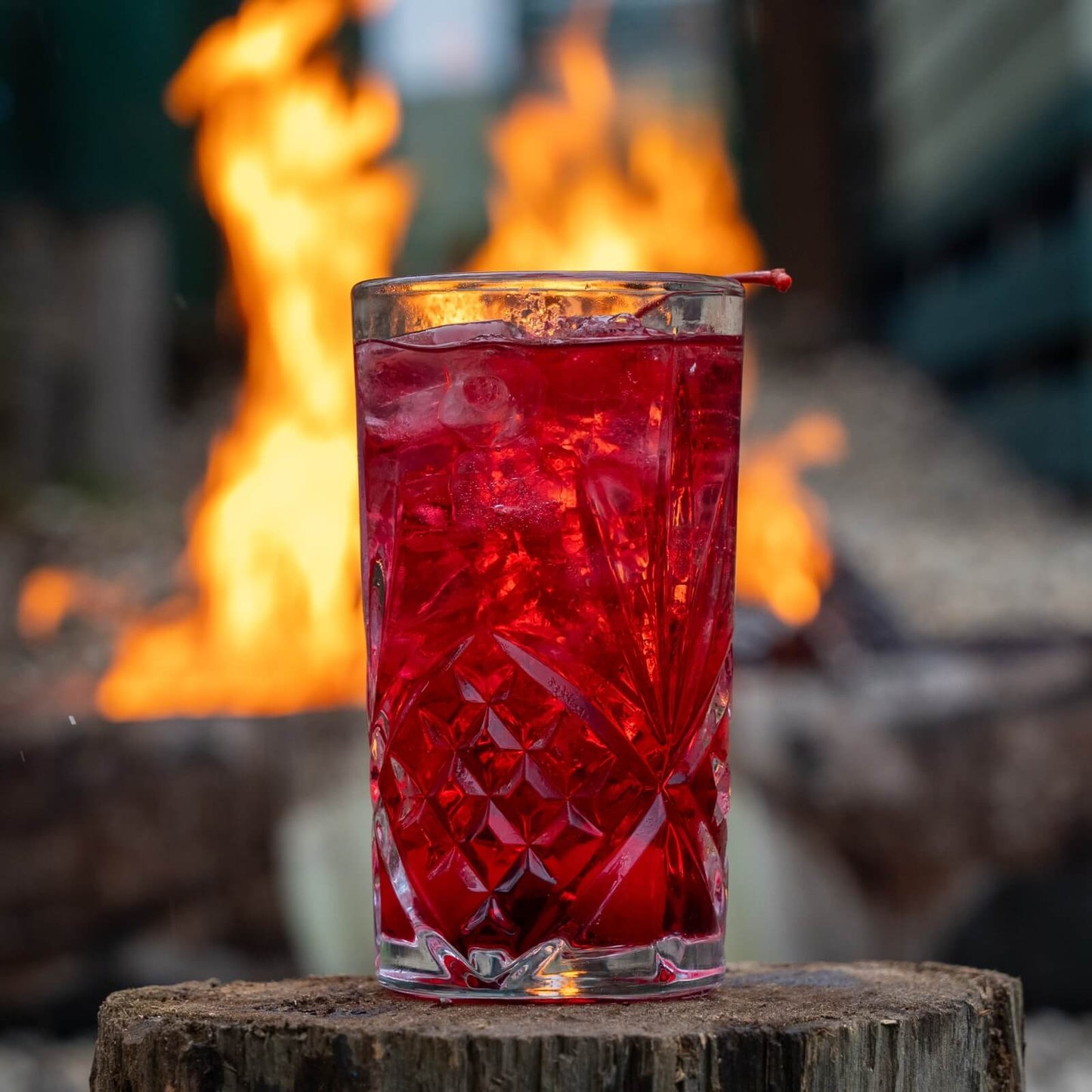 Shirley temple mocktail sitting on a wooden log with blurred out fire pit in the background