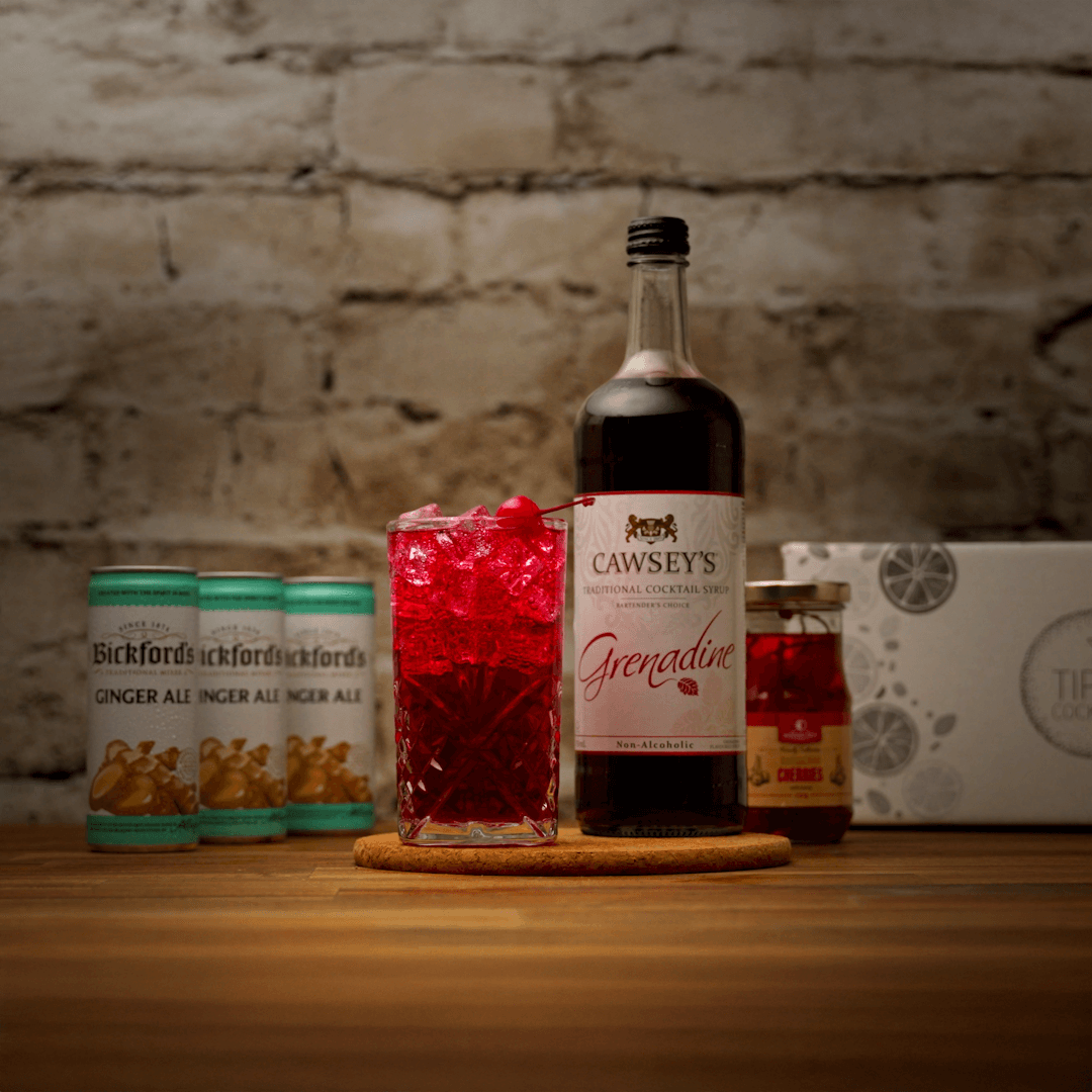 Bottle of Cawsey's Grenadine with a glass of red liquid on a wooden surface against a brick wall.