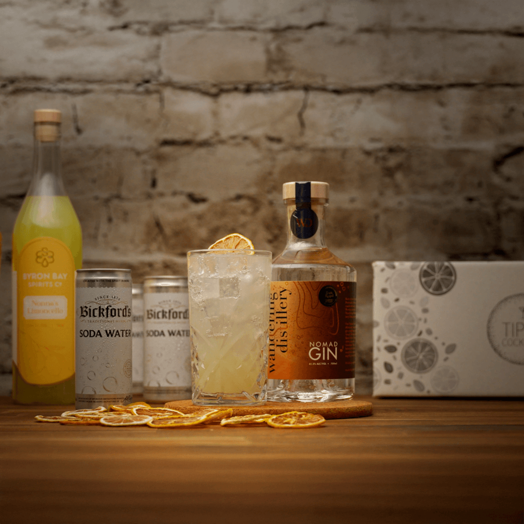summer Zest Cocktail with Wandering Distillery Gin bottle, glass with a drink, and ingredients on a wooden surface with a stone wall background.