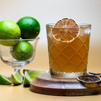 Glass of The Forecast Cocktail drink with dried lime garnish on a wooden coaster, surrounded by limes.