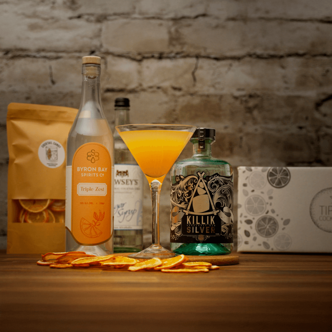 Tiki Orange Daiquiri cocktail kit set up with killik silver rum, byron bay spirit co triple zest, orange slices and sugar syrup on a wooden board against a stone background. 