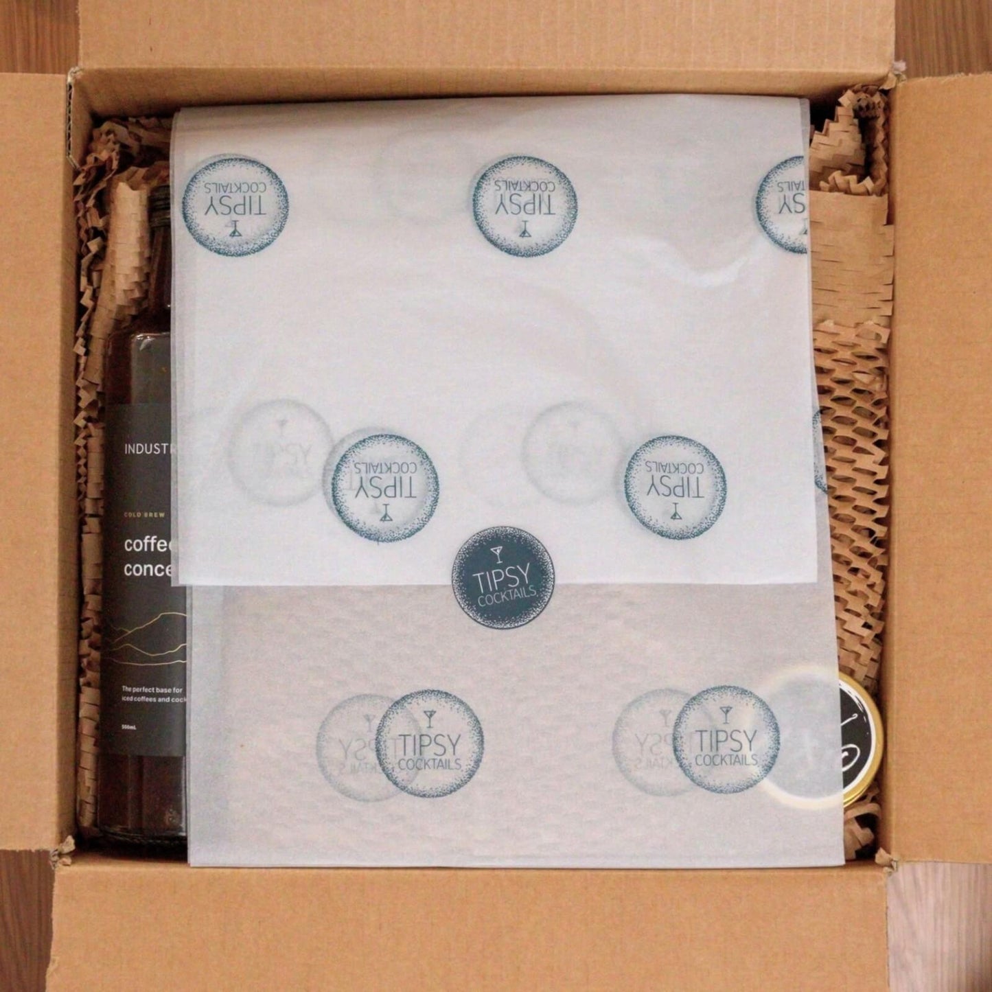 Open Tipsy Cocktails Kit showing wrapping paper sealed with sticker and coffee concentrate on the side in a cardboard box. 