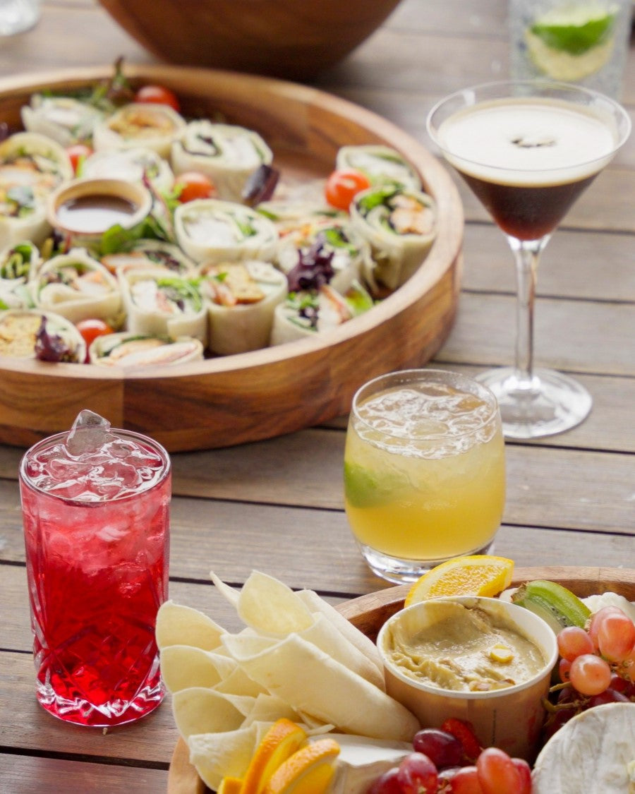 4 cocktails spread out on a wooden outdoor table around lunch platters and serving plates with persons hand reaching for one of the platters.