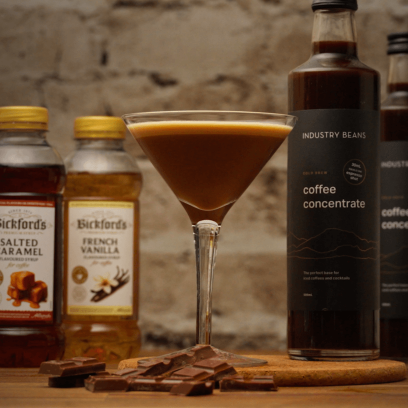 Velvet Hour Coffee Mocktail with chocolate at base of martini glass on a wooden board with a stone background. Mocktail ingredients in background