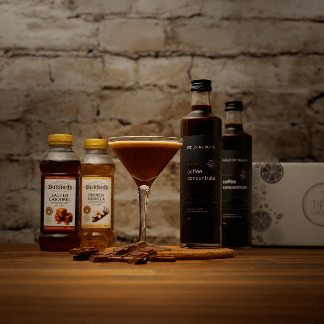 Bottles of Bickford's syrups and Industry Beans coffee concentrate on a wooden surface with a brick wall background. Coffee mocktail sitting in the middle of the ingredients with chocolate at the base of the glass