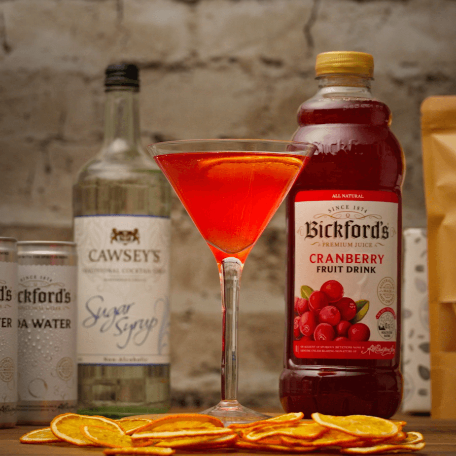 Virgin Cosmopolitan Mocktail with dried orange slices at the base of the glass on a wooden bench. Surrounded by other kit ingredients including cranberry juice and sugar syrup