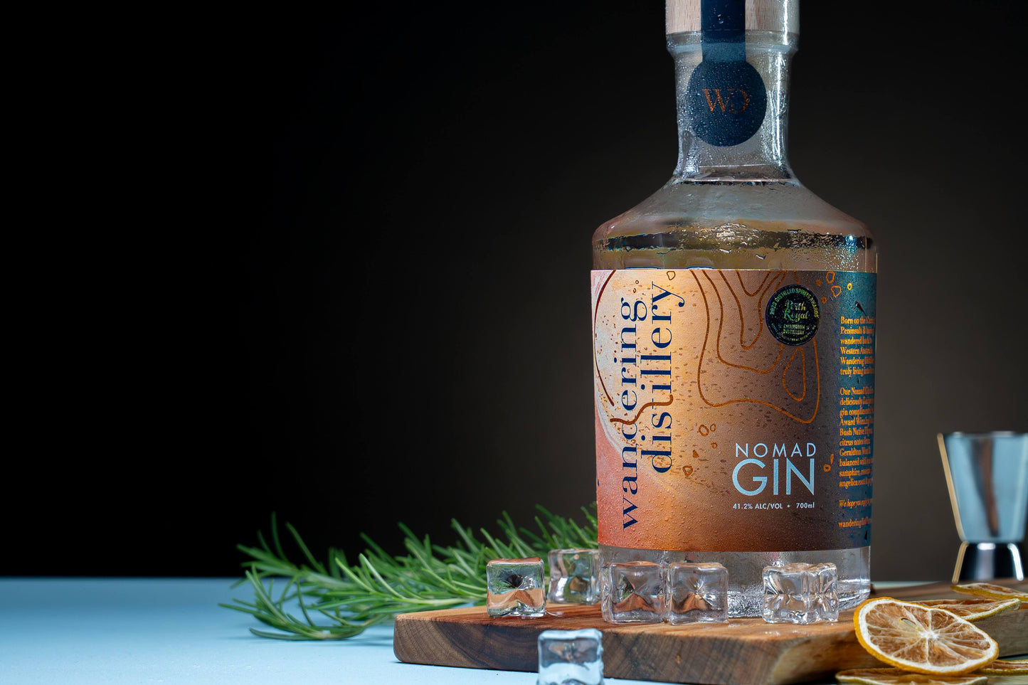 Bottle of Wandering Distillery Nomad Gin on a wooden board with ice, dried lime slices and fresh rosemary. 