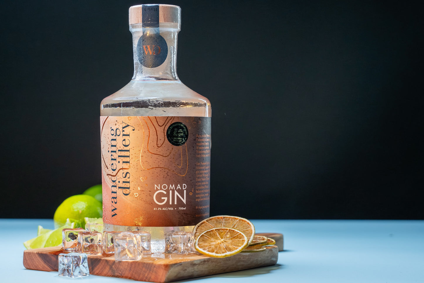 Wandering Distillery Nomad Gin on a wooden board surrounded y ice, dried lime garnishes and fresh lime in the background.