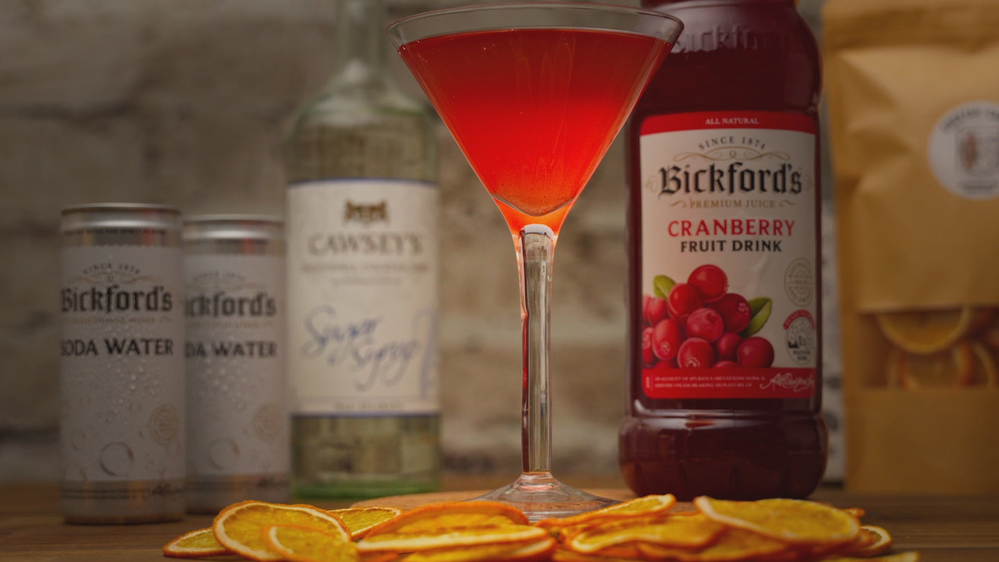 Video shows a close up of a mocktail in a martini glass, zooming out to see mocktail kit contents such as cranberry juice, soda water and other ingredients with a stone background. 