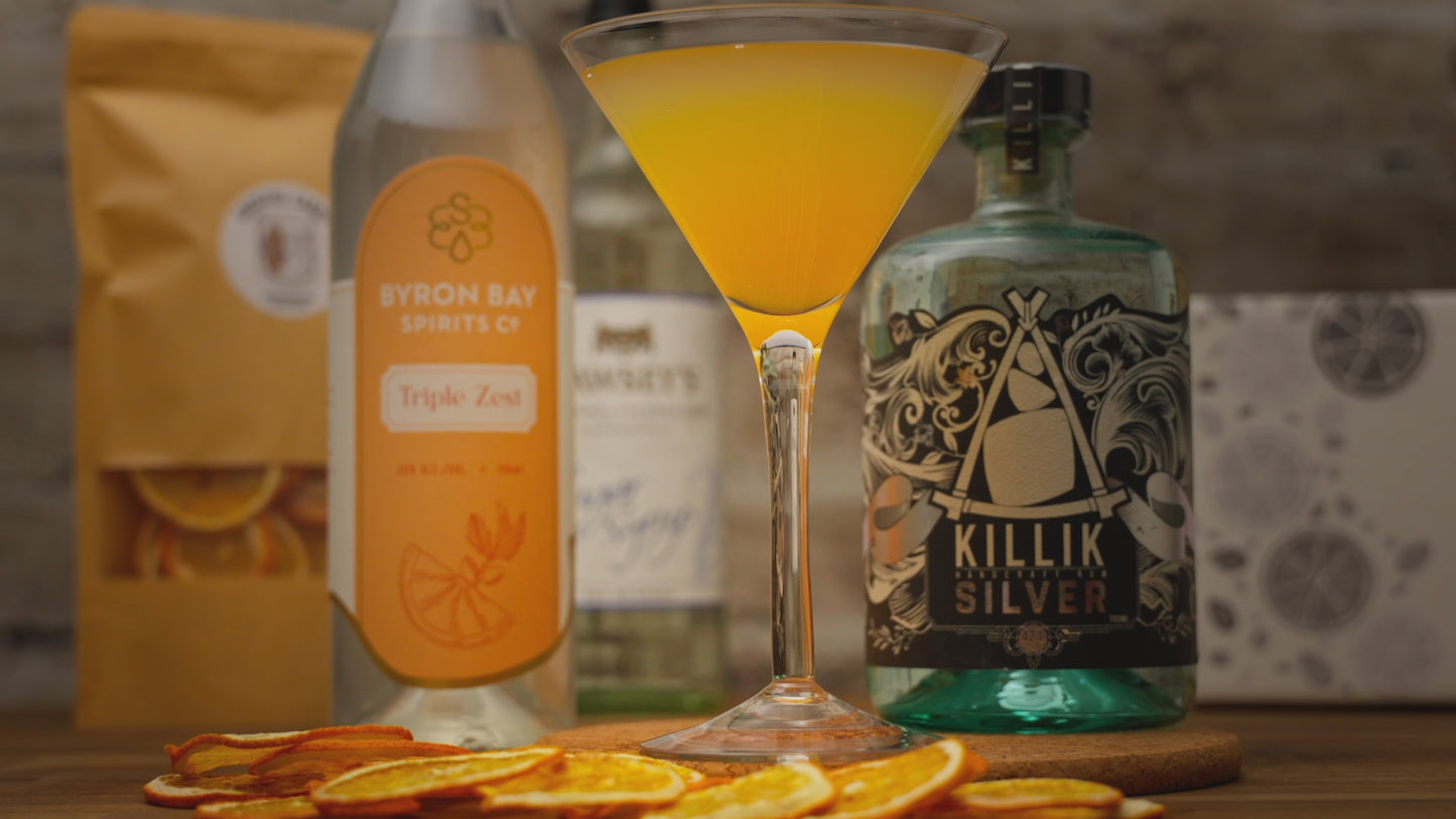Video showing Tiki Orange Daiquiri Cocktail in a tall martini glass, person garnishing with orange slice then zooming out to show cocktail surrounded by kit contents including Killik silver bottle, byron bay triple zest, orange slices and sugar syrup. video zooms back in to show the reverse action of garnishing drink