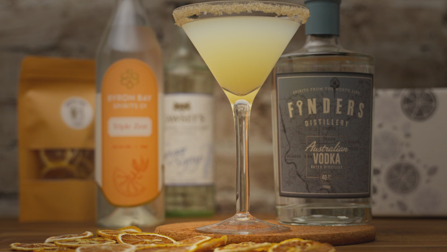 Video of a lemon drop martini being garnished with a dried lemon slice, zooming out to show alcohol bottles included in cocktail kit like Finders Distillery vodka, Byron Bay Spirits Triple Zest and dried garnished. Video zooms back in and reverses the garnish action