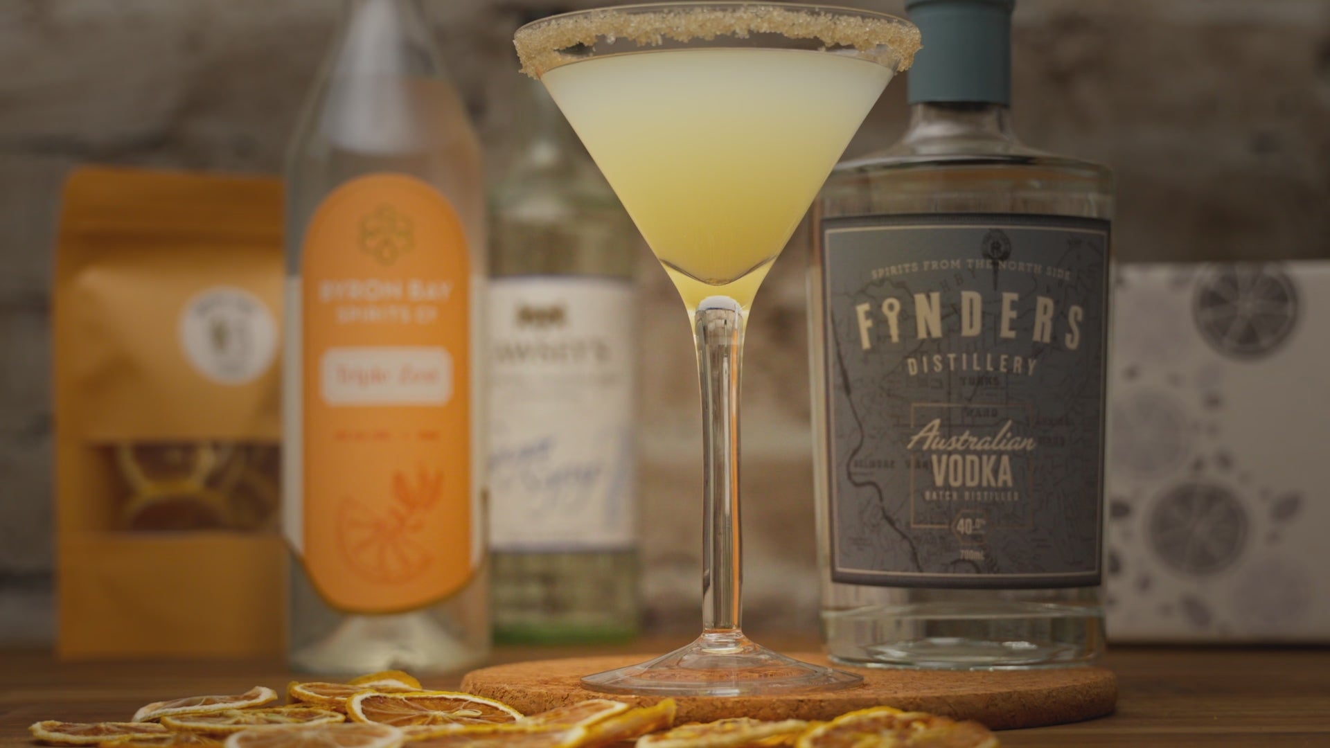 Video of a lemon drop martini being garnished with a dried lemon slice, zooming out to show alcohol bottles included in cocktail kit like Finders Distillery vodka, Byron Bay Spirits Triple Zest and dried garnished. Video zooms back in and reverses the garnish action