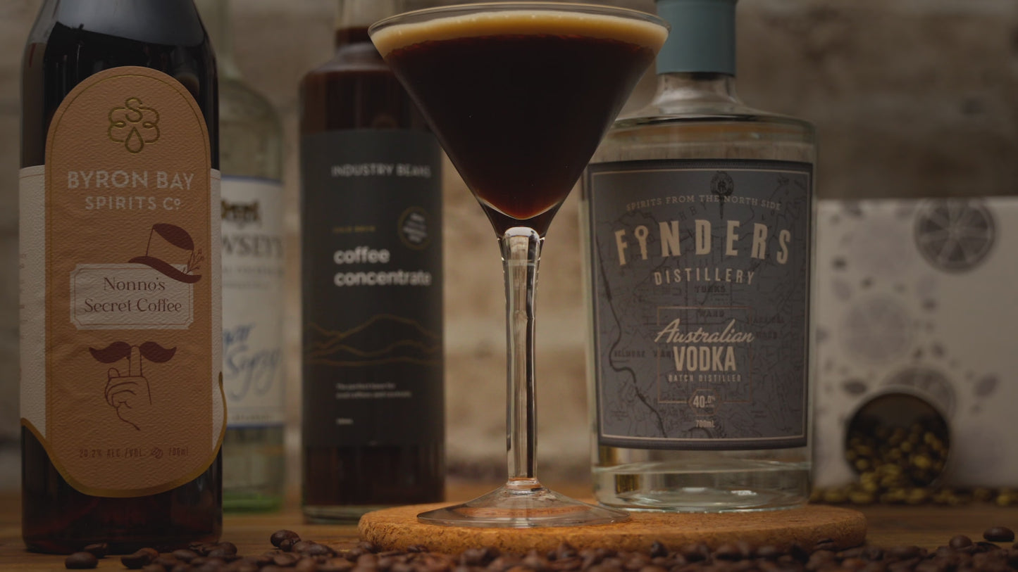 Video of the Espresso Martini Cocktail Kit. Espresso Martini Cocktail in tall martini glass with person garnishing drink with a coffee bean. Video zooms out to show kit contents including Finders Distillery Vodka, Byron Bay Spirit Co's Coffee Liqueur, Industry Beans Coffee Concentrate and sugar syrup with coffee beans surrounding items. Video reverses back in including a reverse of the drink being garnished. 
